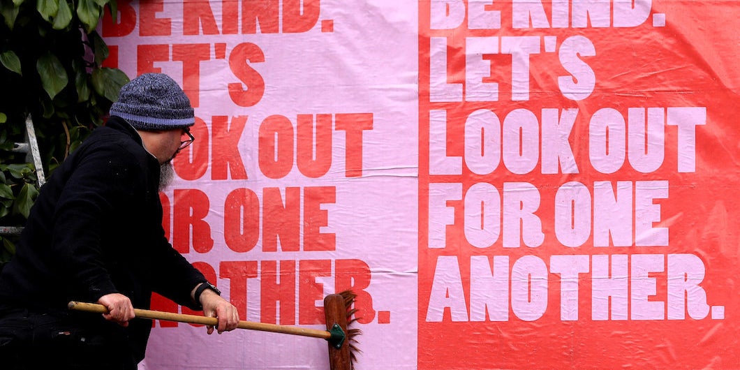 A person pasting a poster to a wall. The poster says: "Be kind. Let's look out for one another."