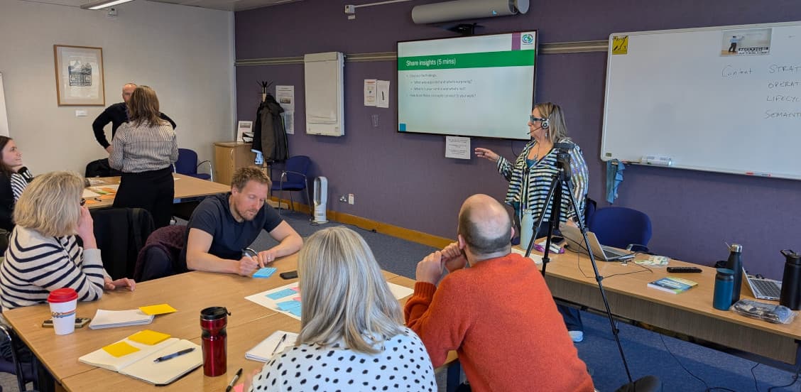 A group of people sitting in a workshop writing on sticky notes. Rahel Anne Bailie stands at the front facilitating the session.